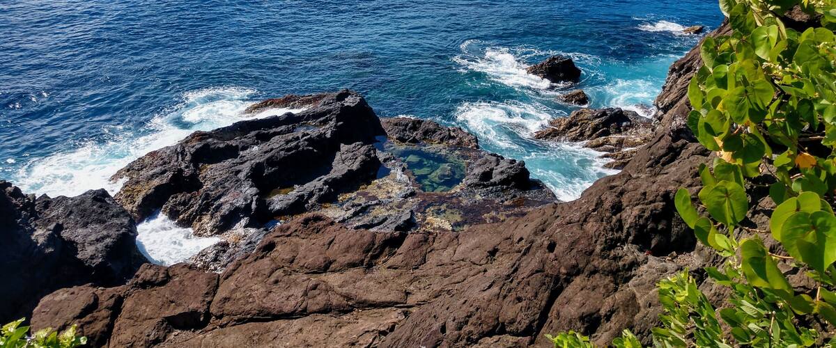 A coastal landscape with rocky shores, clear blue sea, and green hills in the background.