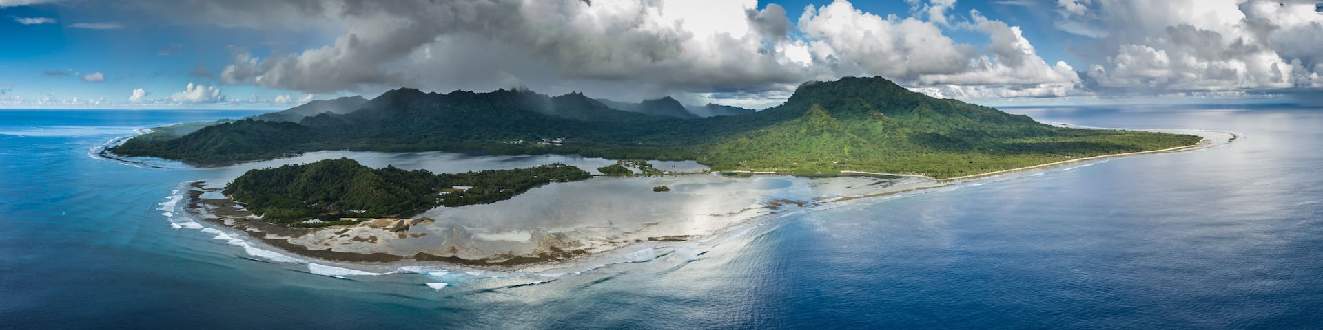 A tropical island with a large mountain, surrounded by clear blue waters and a partly cloudy sky.