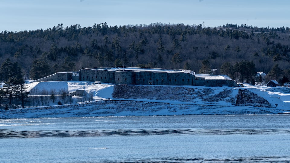 A snow-covered building complex by a frozen lake with a forested hill in the background.