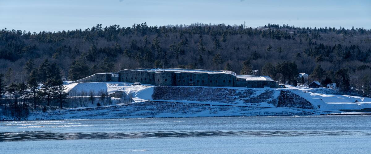 A snow-covered building complex by a frozen lake with a forested hill in the background.