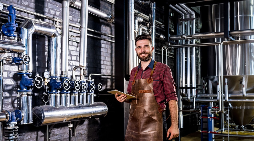Young male brewer in leather apron supervising the process of beer fermentation at modern brewery factory