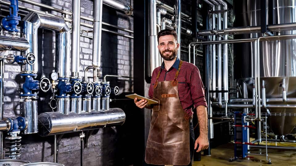 A person in a checkered shirt and leather apron standing in a brewery with stainless steel tanks and pipes.
