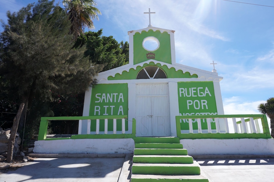 Una pequeña iglesia con fachada verde y blanca, una cruz en la parte superior y las palabras 'Santa Rita' y 'Ruega por nosotros' a los lados.
