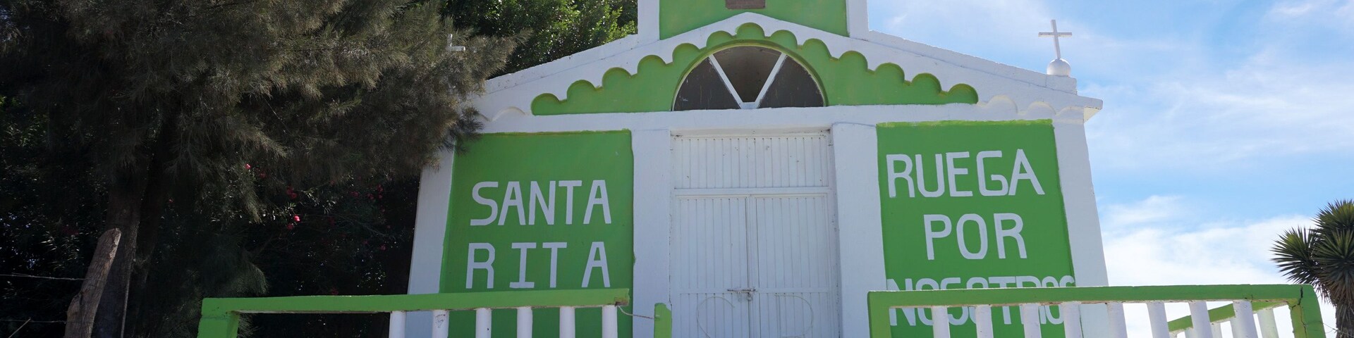 Una pequeña iglesia con fachada verde y blanca, una cruz en la parte superior y las palabras 'Santa Rita' y 'Ruega por nosotros' a los lados.