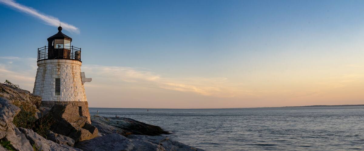 A lighthouse on a rocky coastline with calm sea and a clear sky.