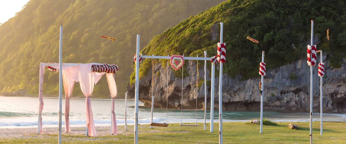 A beach wedding setup with a swing, white columns, and heart-shaped decorations against a backdrop of a cliff and ocean.