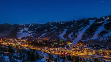 A snow-covered mountain town at night with illuminated buildings and a crescent moon in the sky.