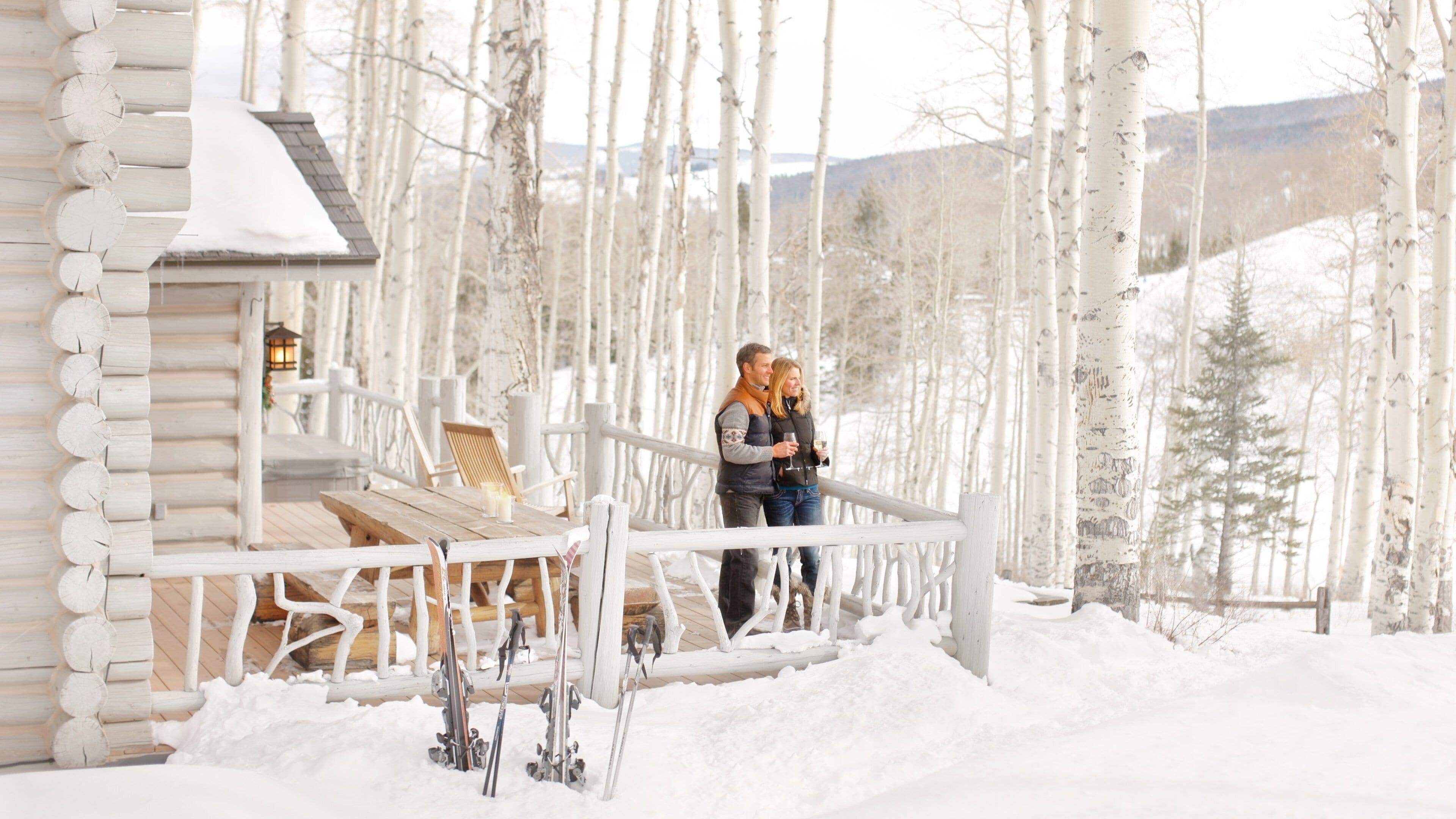 A wooden cabin with a deck, surrounded by snow-covered trees and a snowy landscape.