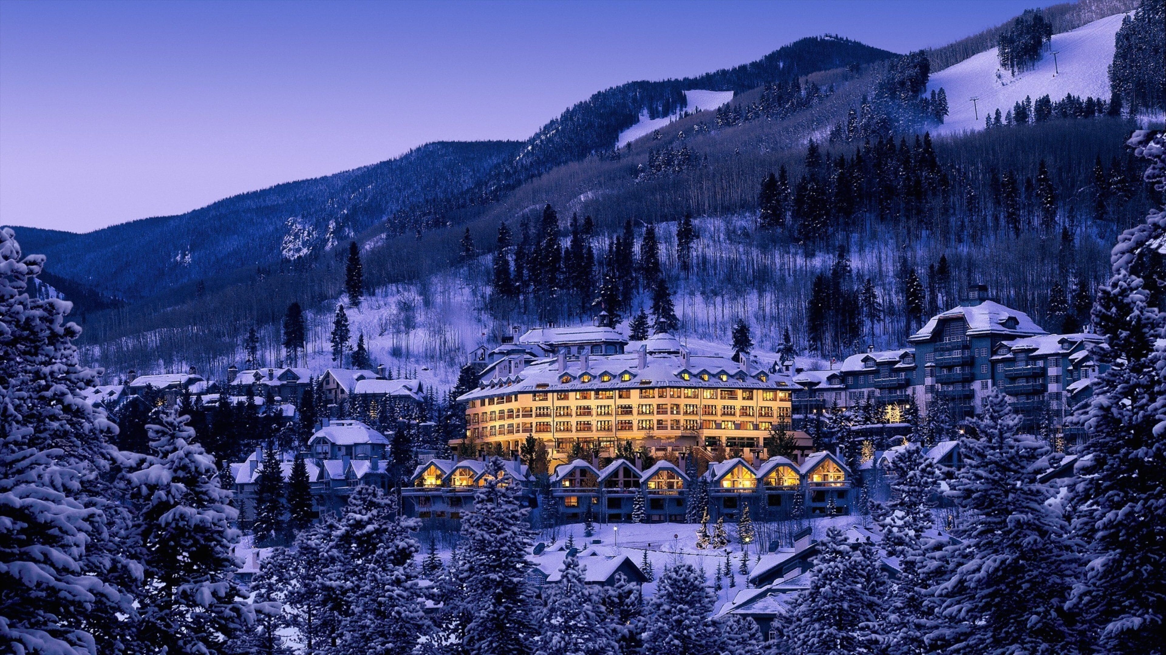 A snow-covered lodge with illuminated windows, surrounded by trees and mountains.