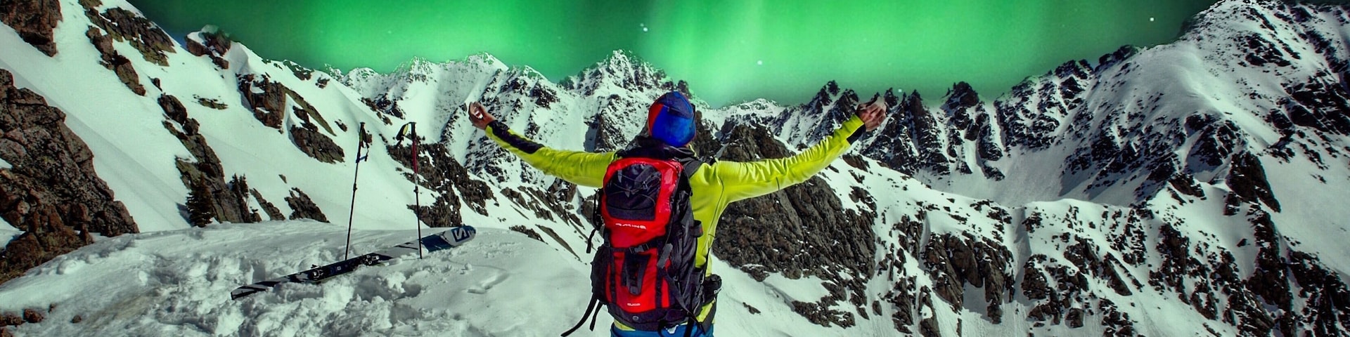 Eine Person steht auf einem verschneiten Berg, die Arme ausgebreitet, und blickt auf ein grünes Polarlicht.