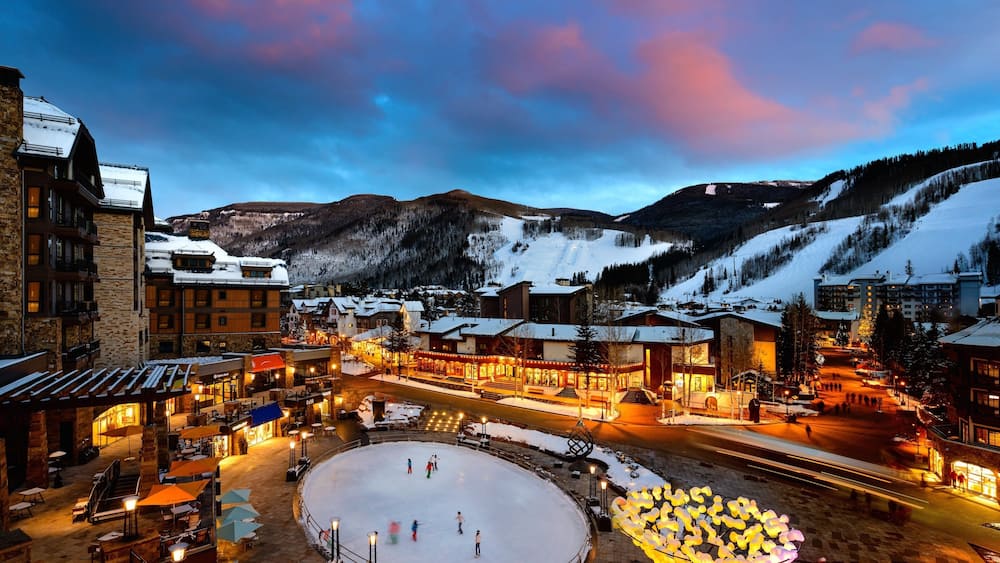 A snow-covered town square with buildings, a frozen pond, and mountains in the background.