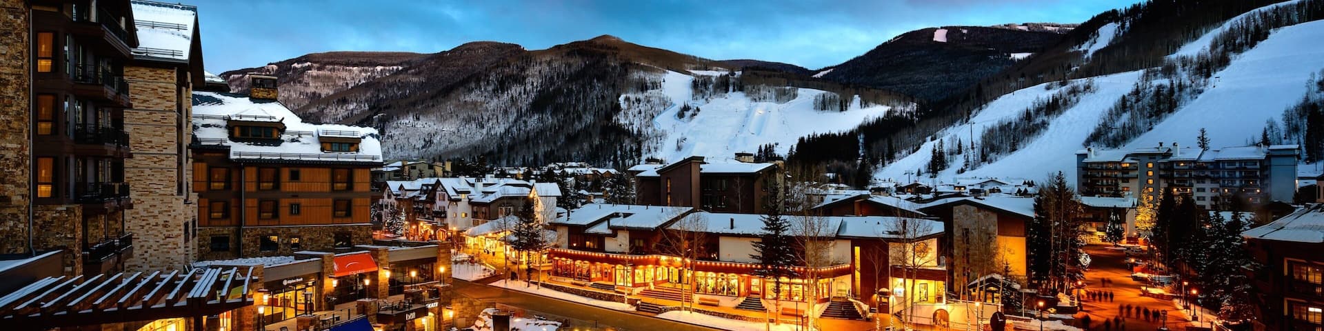 A snow-covered town square with buildings, a frozen pond, and mountains in the background.