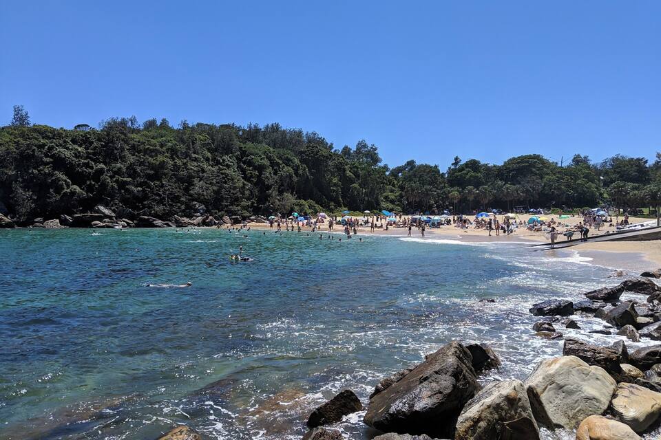 A beach with clear blue water, rocky shoreline, and a forested hill in the background.