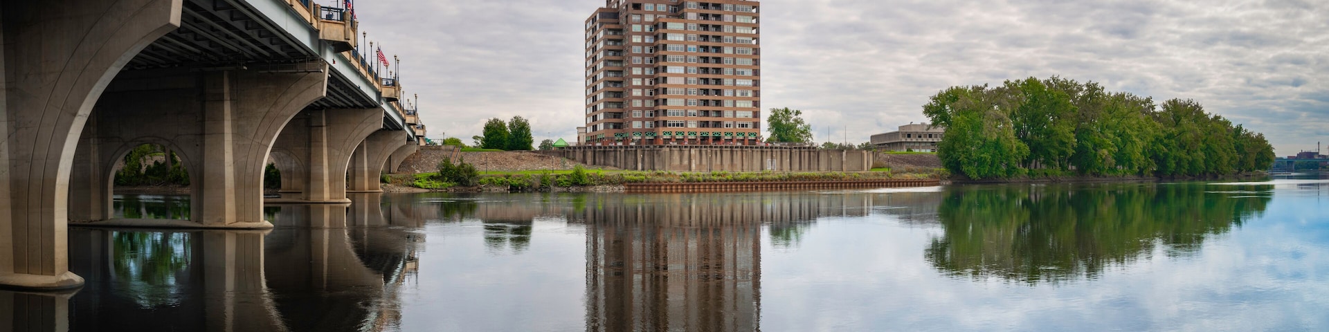Un puente sobre un río tranquilo con su reflejo, un edificio alto con techo verde y árboles a lo largo de la orilla.