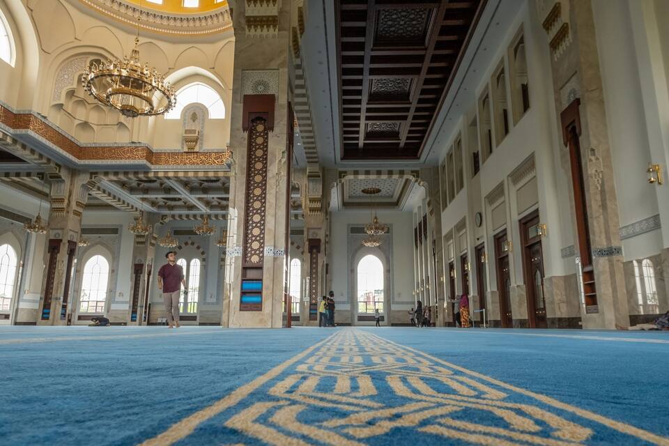 An ornate mosque interior with blue carpet, arched windows, and intricate wall designs.