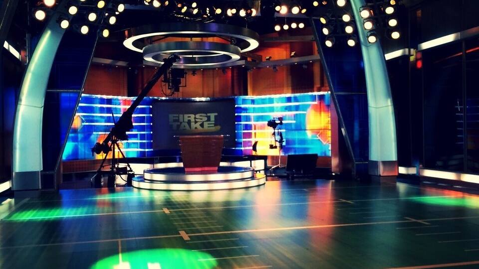 A brightly lit studio with a stage, a large screen displaying 'FIRST TAKE', and a circular platform in the center.