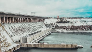 A large dam with a spillway, a river below, and a snowy landscape with buildings in the background.