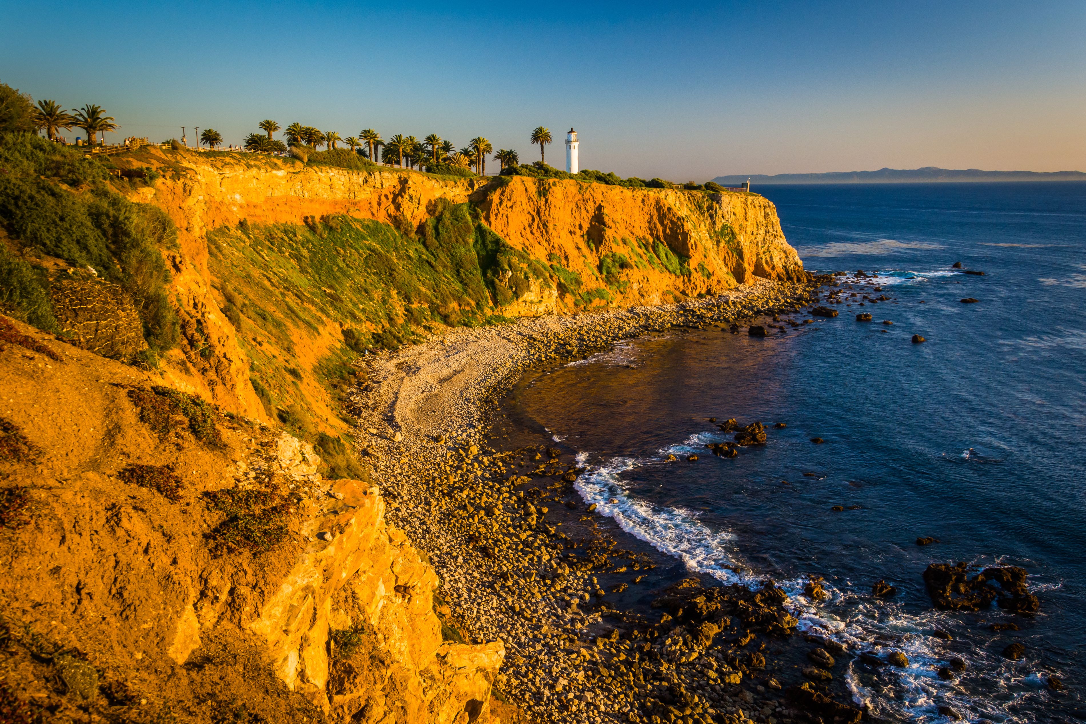 A coastal landscape with a rocky cliff, a lighthouse, and palm trees.