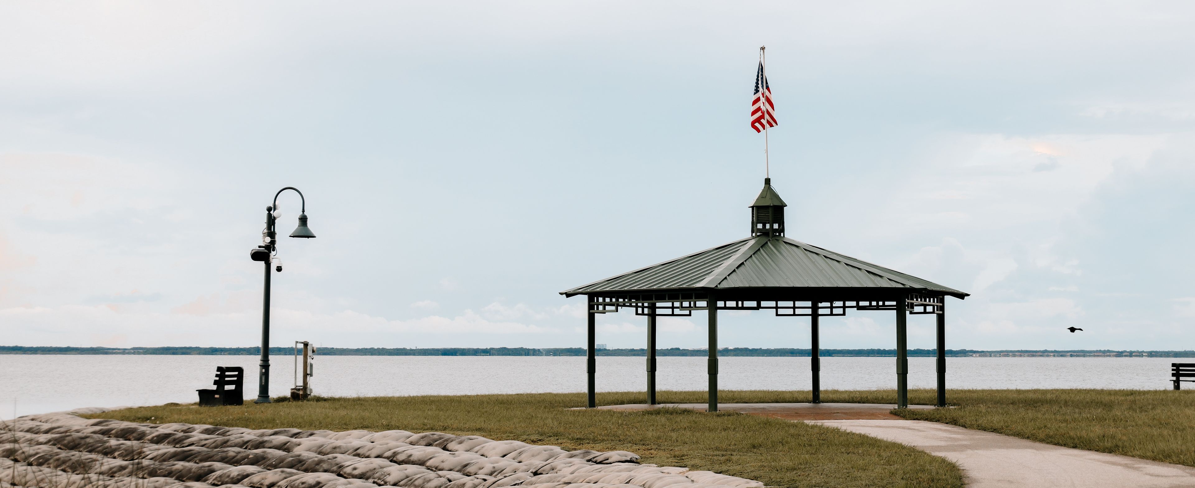 A gazebo with an American flag, a lamp post, and a bench by a lake.