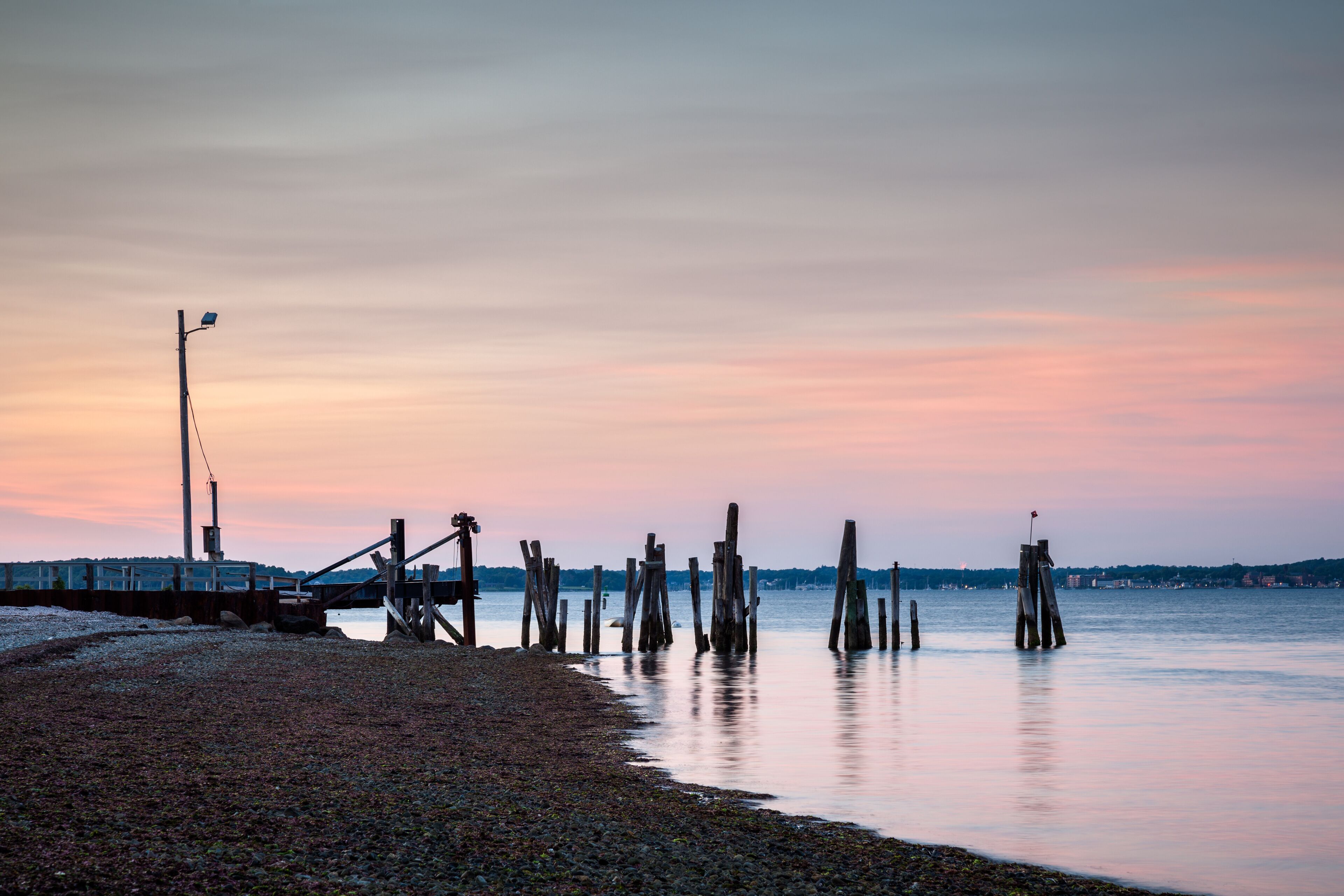 A pebbled beach with wooden posts in the water and a distant city skyline at sunset.