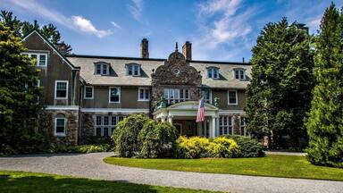 A large, historic mansion with a prominent stone facade, multiple chimneys, and a well-manicured lawn.