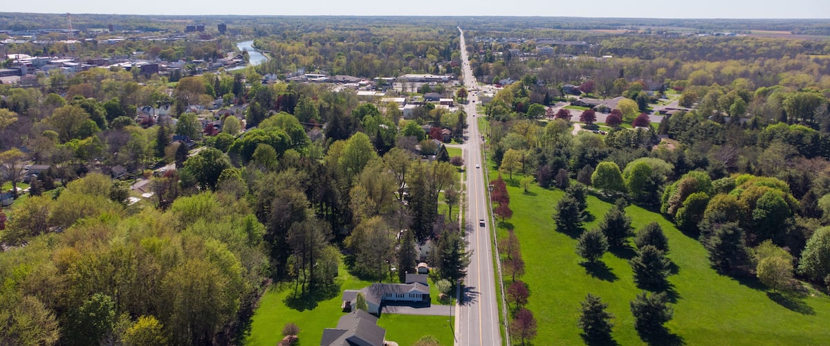 An aerial view of a suburban area with a main road, residential houses, and surrounding greenery.