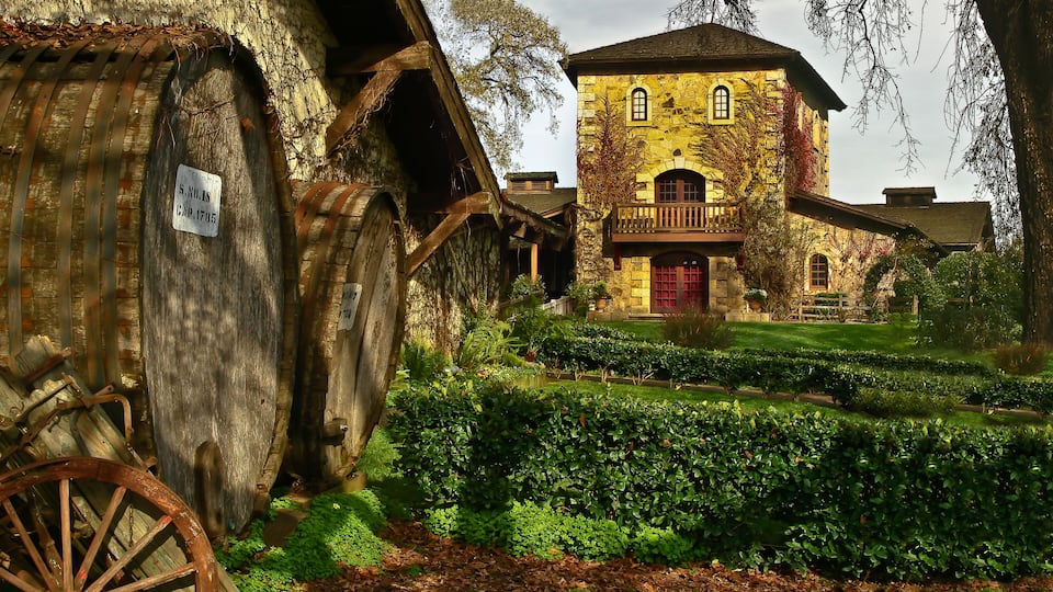 A rustic stone building with a red door and a wooden balcony, surrounded by lush greenery and a vintage wooden cart.