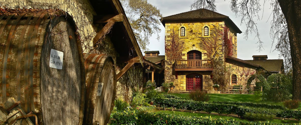 A rustic stone building with a red door and a wooden balcony, surrounded by lush greenery and a vintage wooden cart.