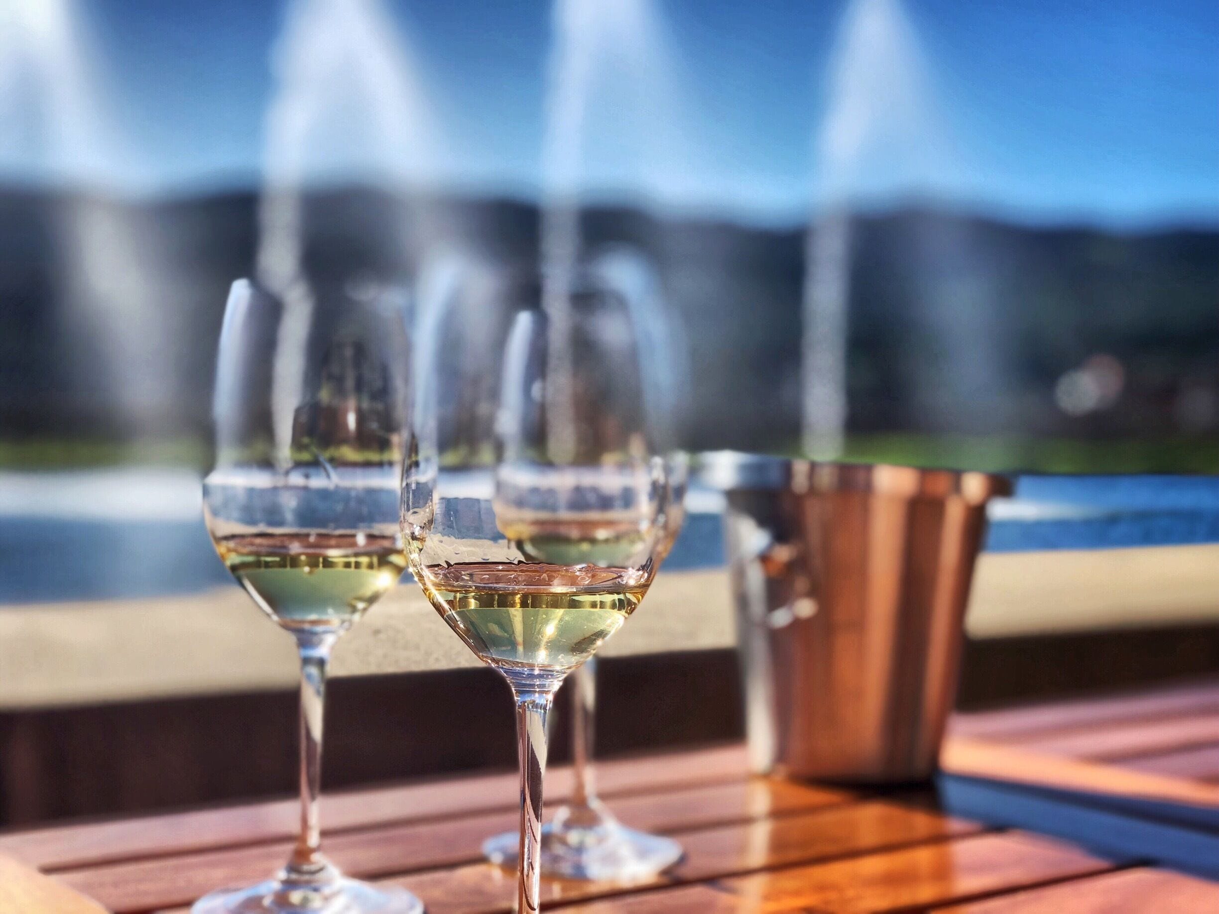 Three wine glasses on a wooden table with a metal bucket, set against a blurred background of a fountain and a mountainous landscape.