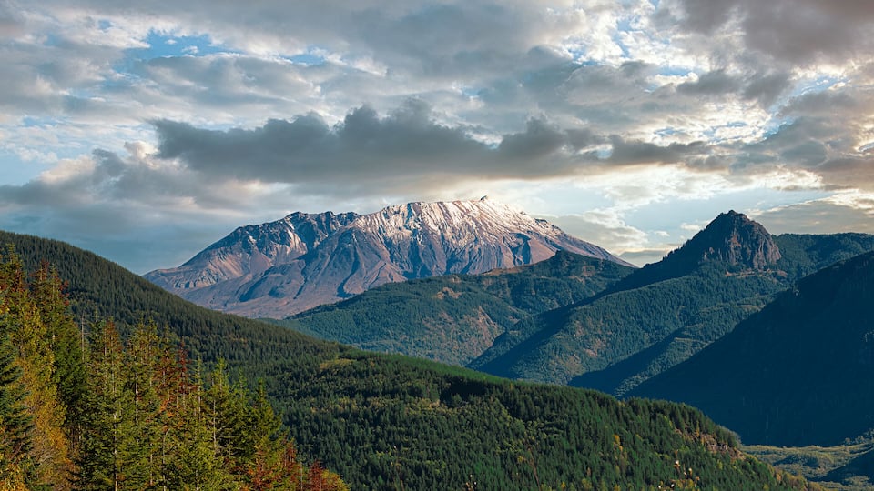 A mountain range with a prominent peak, dense forested valleys, and a cloudy sky.