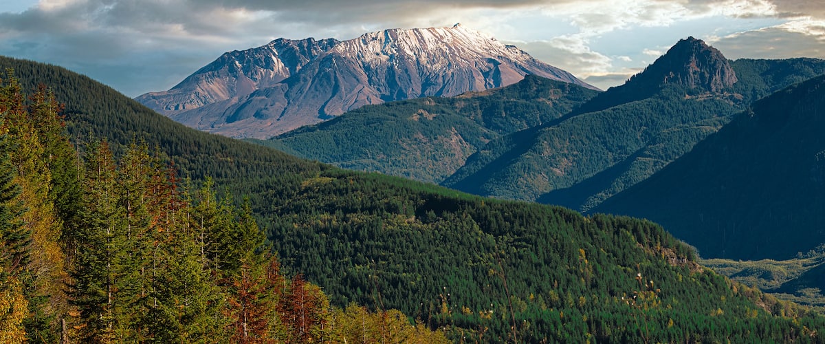 A mountain range with a prominent peak, dense forested valleys, and a cloudy sky.