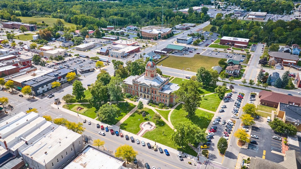An aerial view of a town with a prominent red-brick building with a dome, surrounded by green spaces and a parking lot.