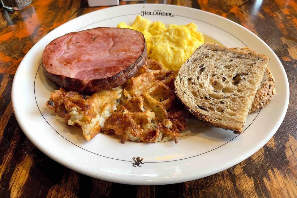 A plate with ham, scrambled eggs, hash browns, and a slice of bread on a Hollander's branded plate.