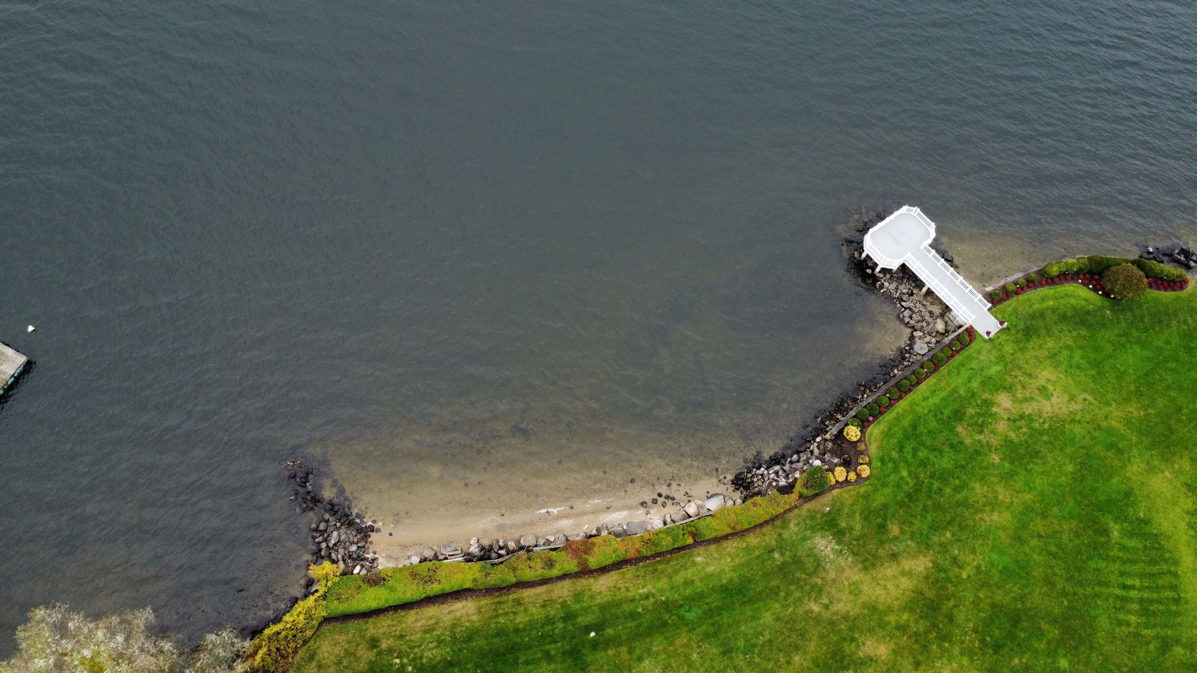 An aerial view of a coastal area with a white pier extending into the water, a grassy embankment, and a rocky shoreline.
