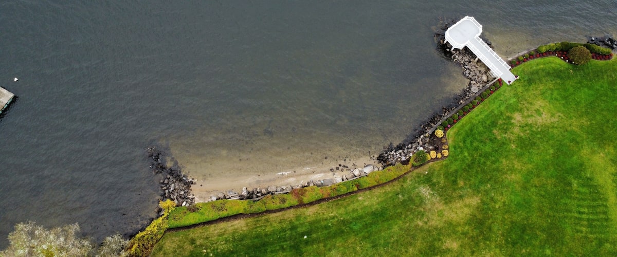 An aerial view of a coastal area with a white pier extending into the water, a grassy embankment, and a rocky shoreline.