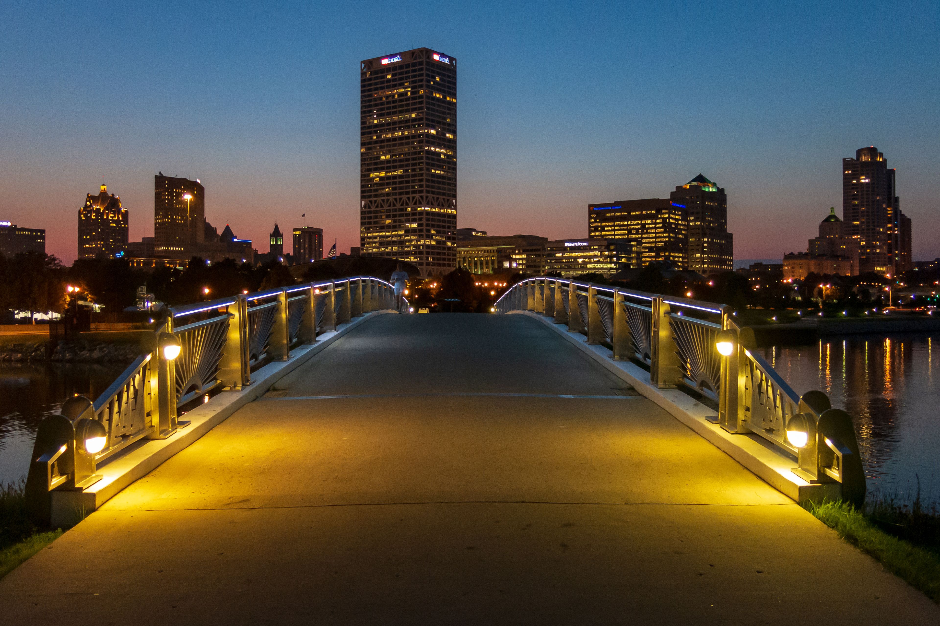 Un puente con barandillas iluminadas que conduce a una silueta urbana al atardecer.
