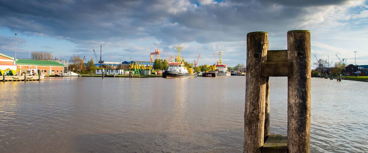 A river with a wooden structure in the foreground and industrial cranes in the background.