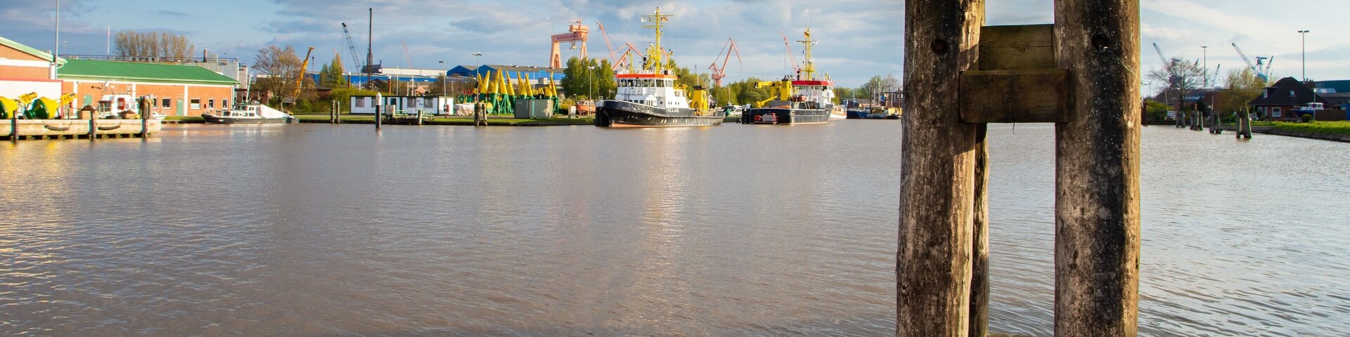 A river with a wooden structure in the foreground and industrial cranes in the background.