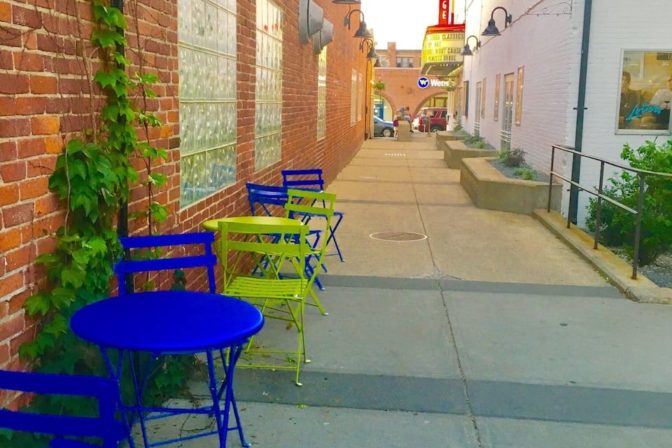 A narrow alley with brick buildings, colorful outdoor seating, and a brightly lit sign.