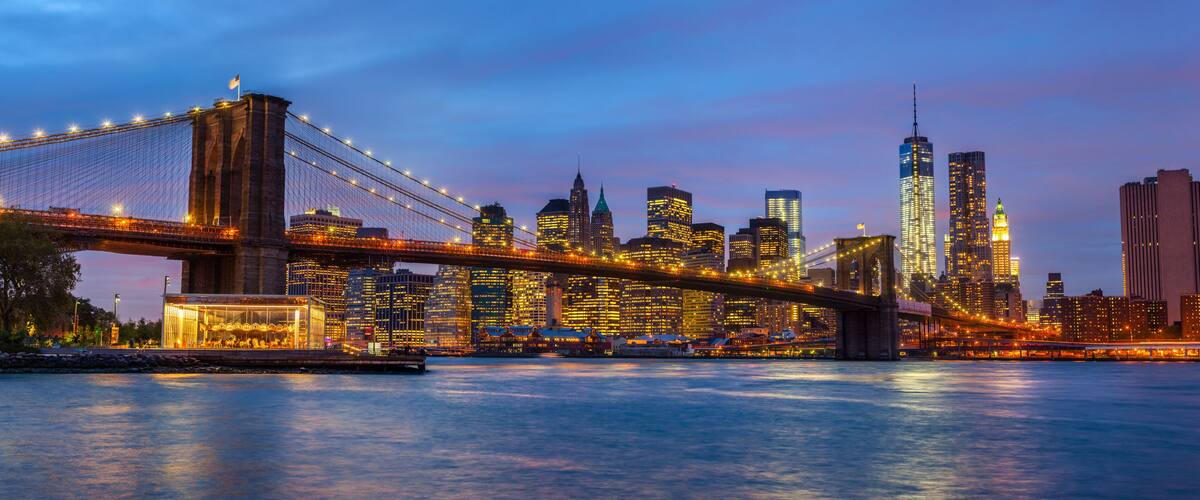 The Brooklyn Bridge at dusk with illuminated buildings in the Manhattan skyline.