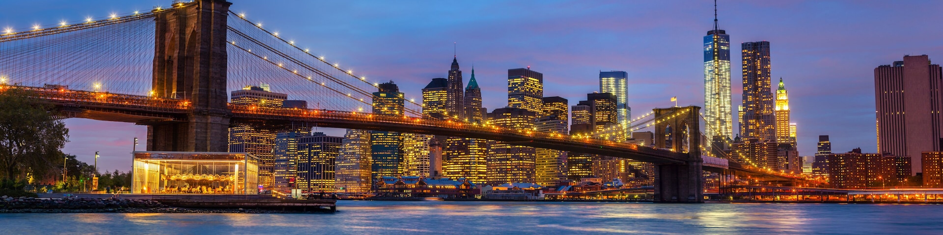 The Brooklyn Bridge at dusk with illuminated buildings in the Manhattan skyline.