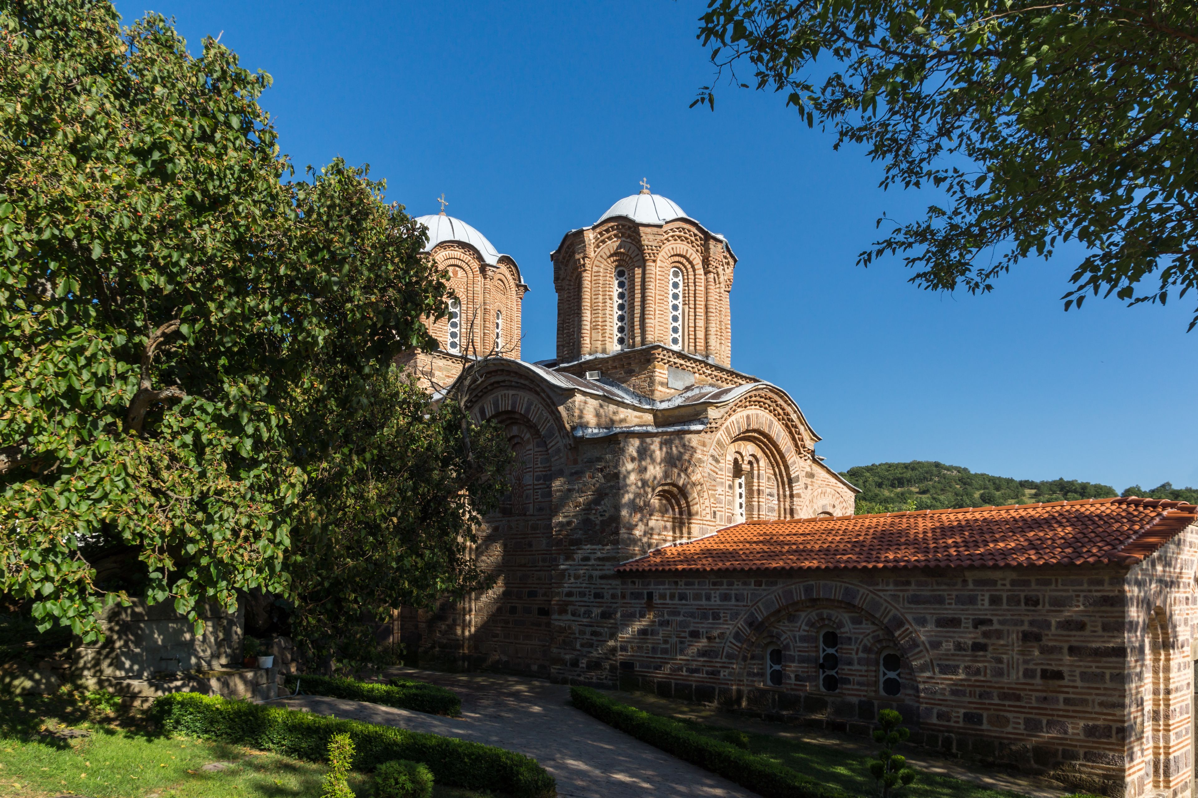 A historic church with two domes and a red-tiled roof, surrounded by greenery and a clear blue sky.