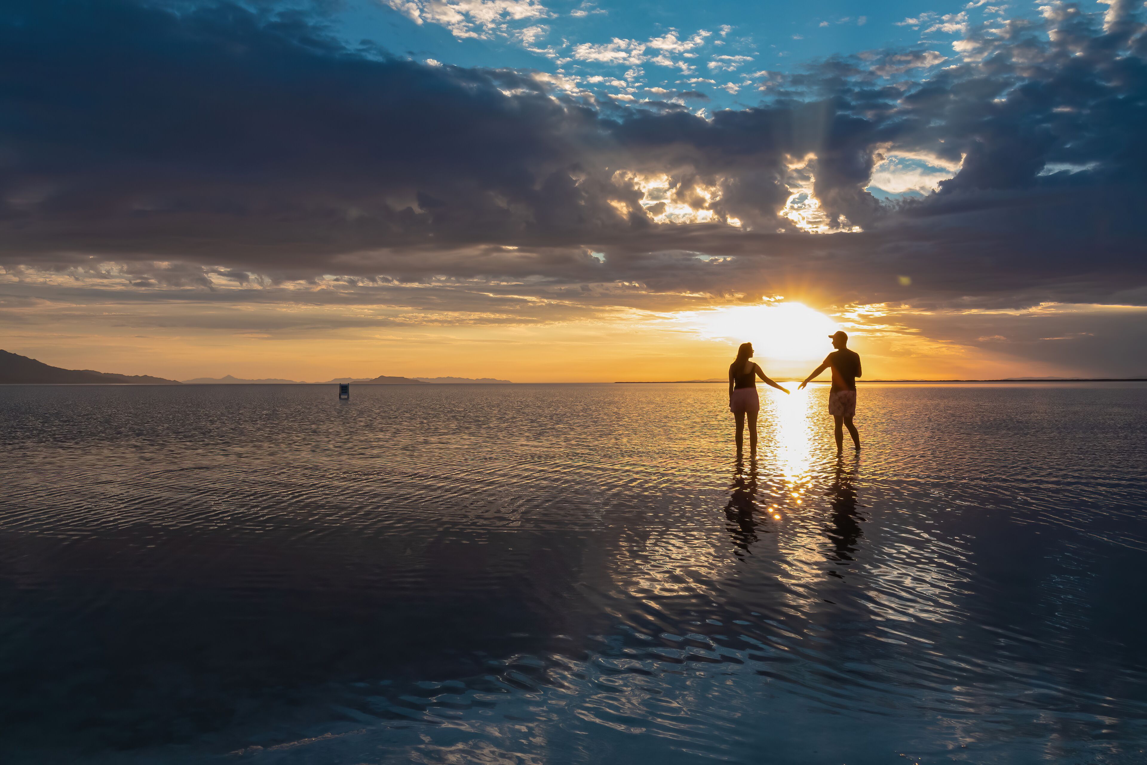 Two people holding hands on a reflective water surface with a sunset in the background.