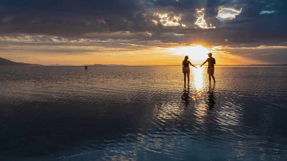 Two people holding hands on a reflective water surface with a sunset in the background.