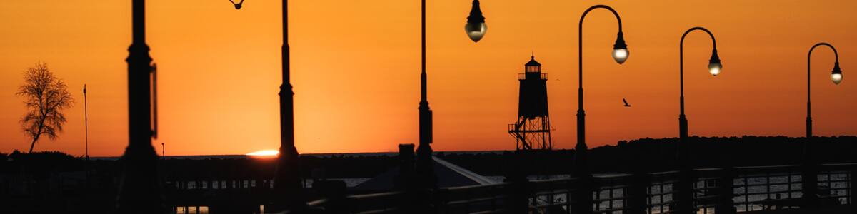 A sunset with street lamps silhouetted against an orange sky, a pier extending into the water, and a lighthouse in the distance.