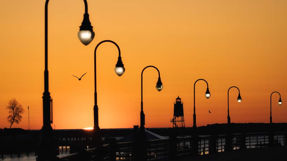 A sunset with street lamps silhouetted against an orange sky, a pier extending into the water, and a lighthouse in the distance.