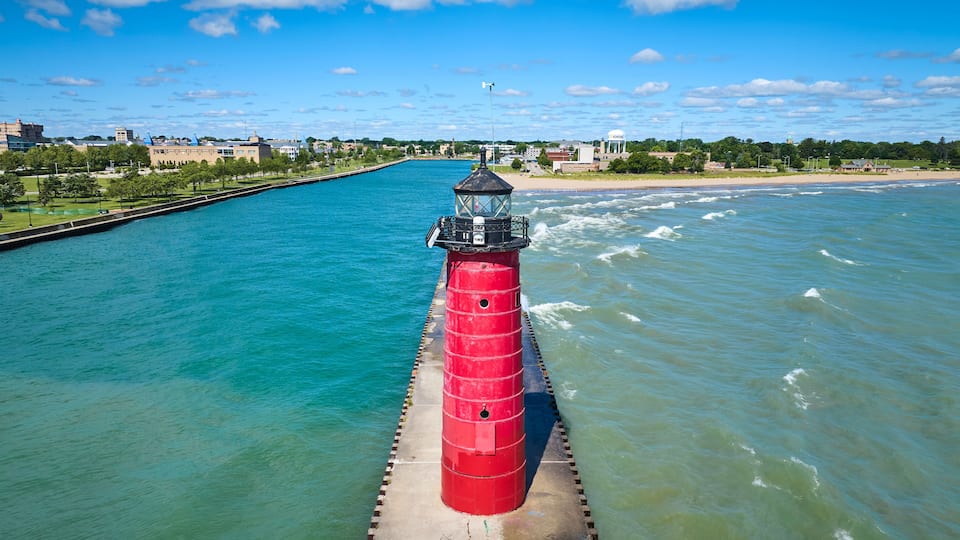 A red lighthouse on a pier extending into a body of water, with a clear sky and a distant shoreline with buildings.