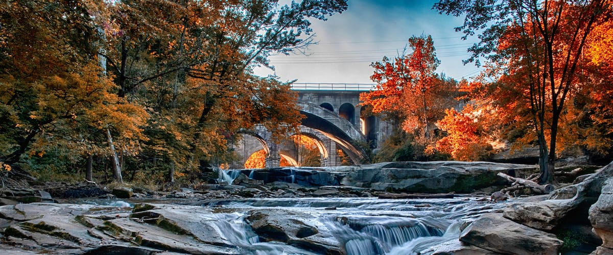 A waterfall cascading over rocks, a stone bridge, and autumn-colored trees.