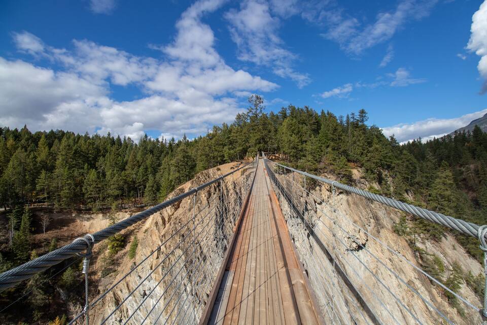 Uma ponte suspensa com tabuleiro de madeira, emoldurada por uma paisagem montanhosa coberta por floresta.