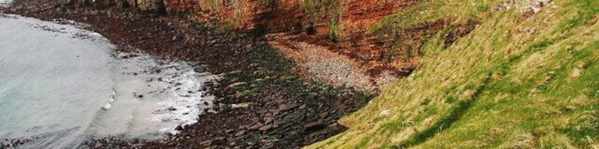 A coastal cliff with red and green rock layers, a grassy slope, and a rocky beach.